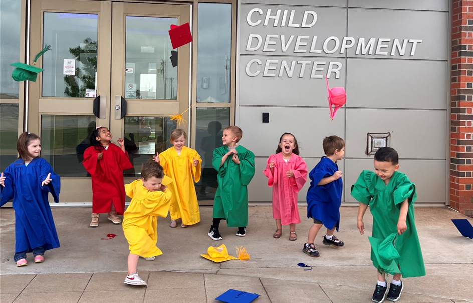 preschool students in graduation gowns throw hats in front of CDC building.